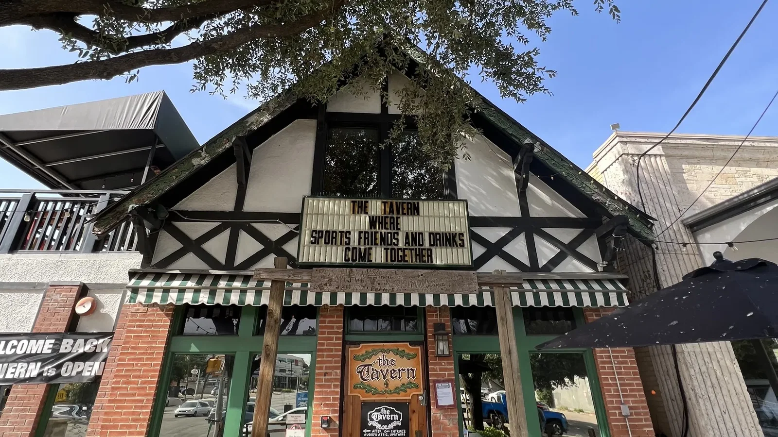 Brick building with Tudor black-and-white trim and green-framed windows sits under a large leafy tree. A striped green awning covers the storefront; a marquee reads, 