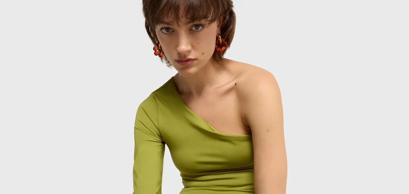 Robe verte asymétrique à manches longues, design ajusté, tissu texturé, accentuée par une ceinture multicolore et des talons roses.