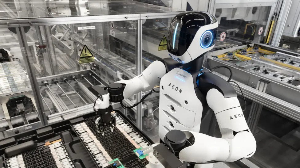 A white and black humanoid robot has a vertical line of blue light down its black plastic face and blue circles where its ears would be. It is working in a factory on what appears to be a production line.
