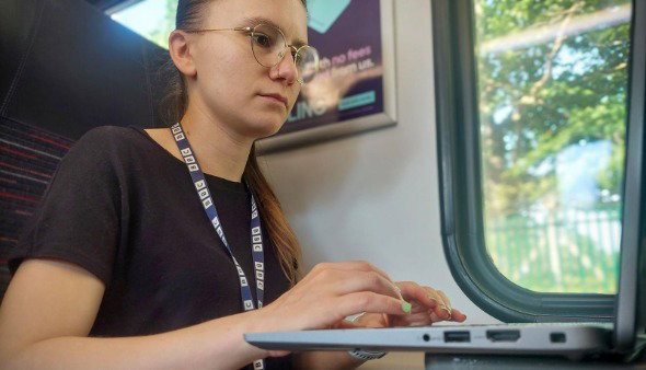 Reporter Grace Dean wears a BBC blue lanyard and taps on a laptop, checking wi-fi access as she travels on a train near a window.