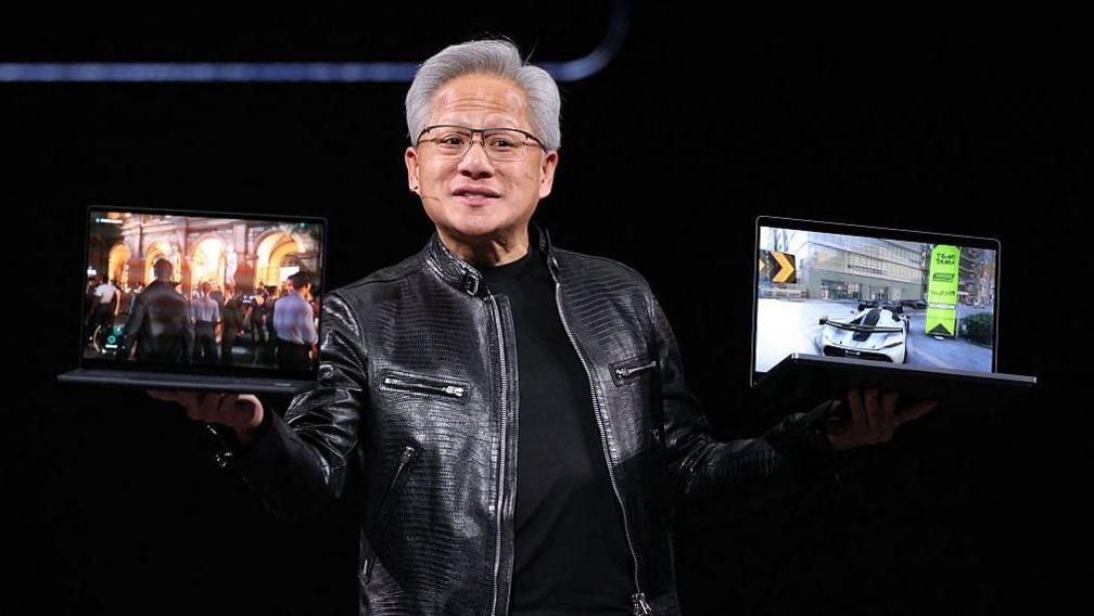 Nvidia's chief executive Jensen Huang holds up two laptop computers which show different gaming displays on a stage. He wears a black leather jacket and glasses.