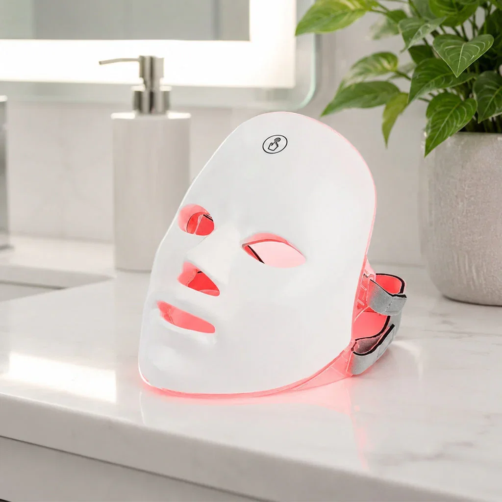LED light therapy mask with red light activation displayed on bathroom countertop next to skincare products and plants.