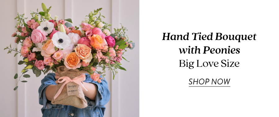 Product feature for the Hand Tied Bouquet with Peonies in Big Love Size, showing a larger burlap-wrapped bouquet with pink, peach, coral, white, and green flowers held in front of a person wearing a blue shirt. Text on the right reads “Hand Tied Bouquet with Peonies, Big Love Size” with a “Shop Now” link.