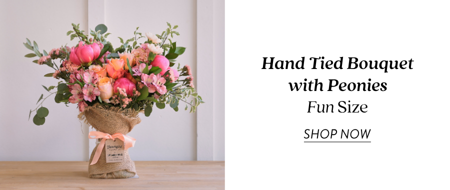 Product feature for the Hand Tied Bouquet with Peonies in Fun Size, showing a colorful burlap-wrapped bouquet with pink, coral, peach, and green flowers on a tabletop. Text on the right reads “Hand Tied Bouquet with Peonies, Fun Size” with a “Shop Now” link.
