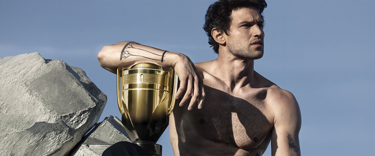 Ein muskulöser Mann mit Tätowierungen posiert neben einem goldenen Pokal vor einem strahlend blauen Himmel.