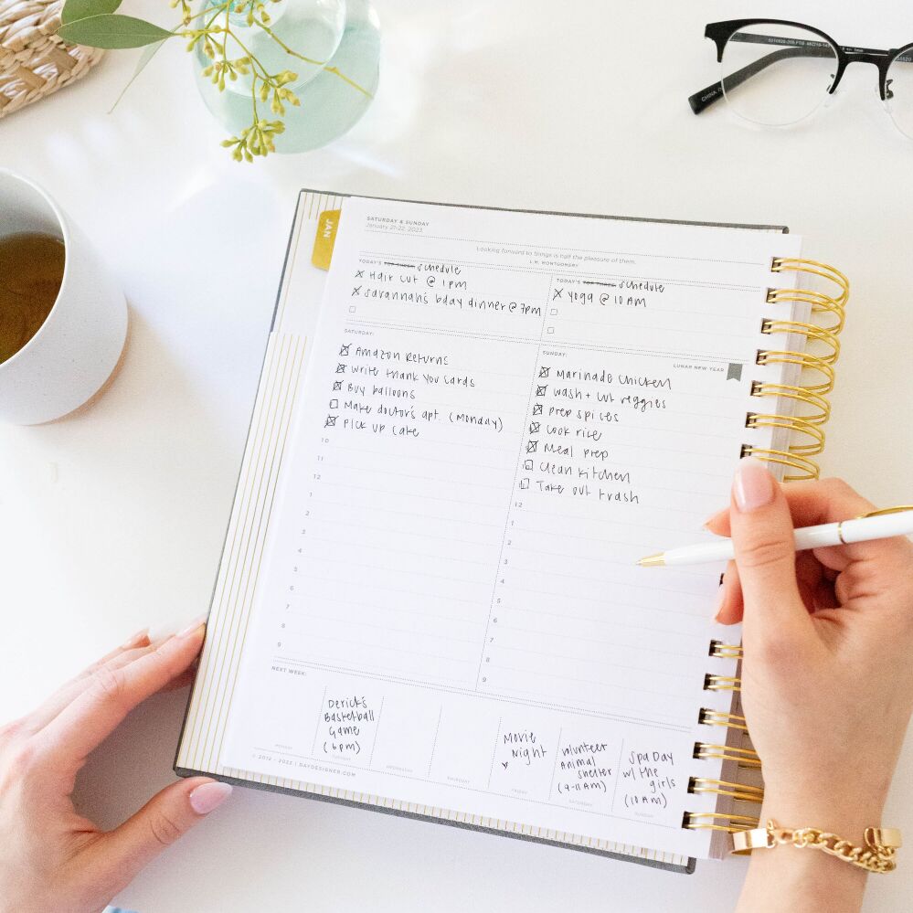 hands holding folded, open book day designer planner, showing written on page, vase and flowers, white background, tea cup, glasses