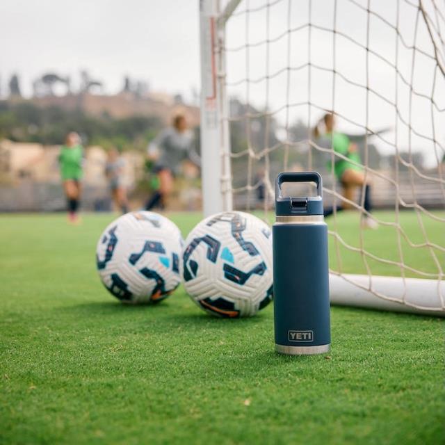 A Yeti bottle and two soccer balls on grass near a goalpost.