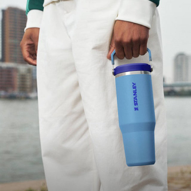 Person in white outfit holding a blue Stanley water bottle near a waterfront. City buildings are in the background.