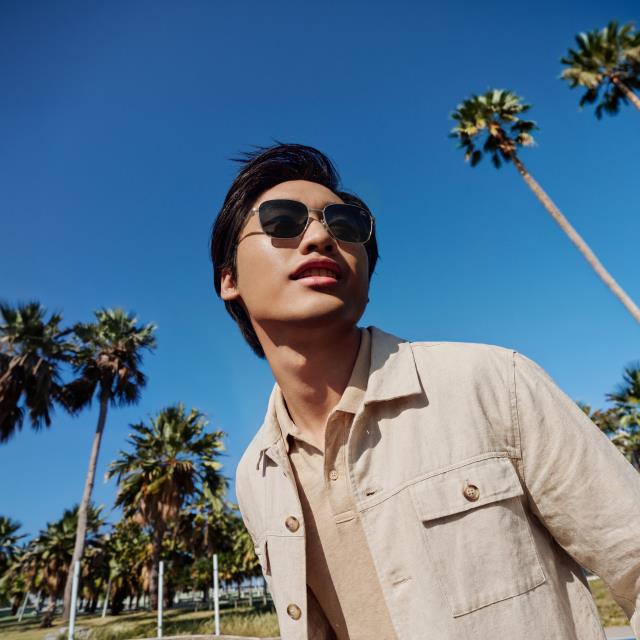 A person wearing sunglasses and a beige jacket stands under a clear blue sky, surrounded by palm trees.