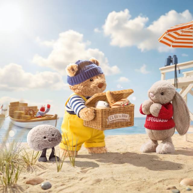 Stuffed animals dressed as a sailor and lifeguard at a beach, with beach umbrellas and a boat in the background.