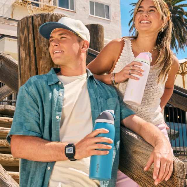 Two people smiling outdoors, holding reusable water bottles. They are casually dressed, with a wooden structure and palm trees in the background.