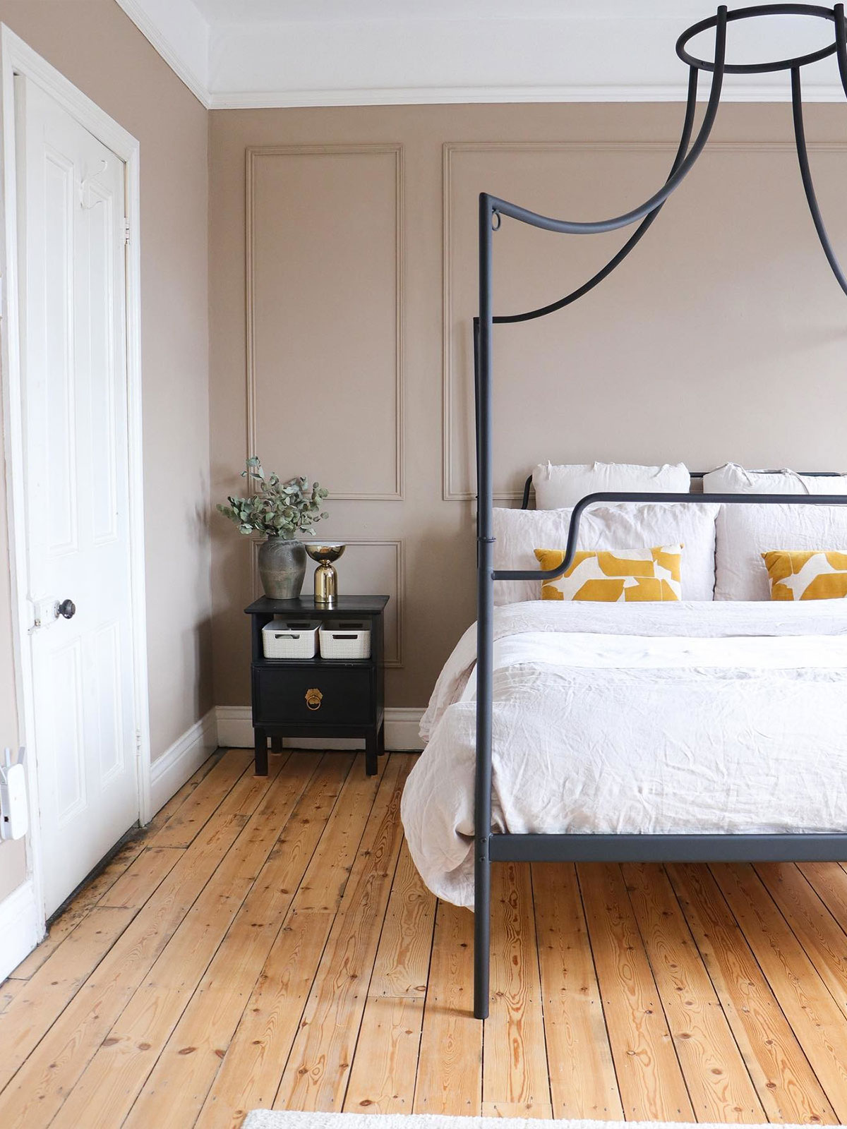 A calm bedroom with a black four-poster bed, mustard cushions, and wooden floors.