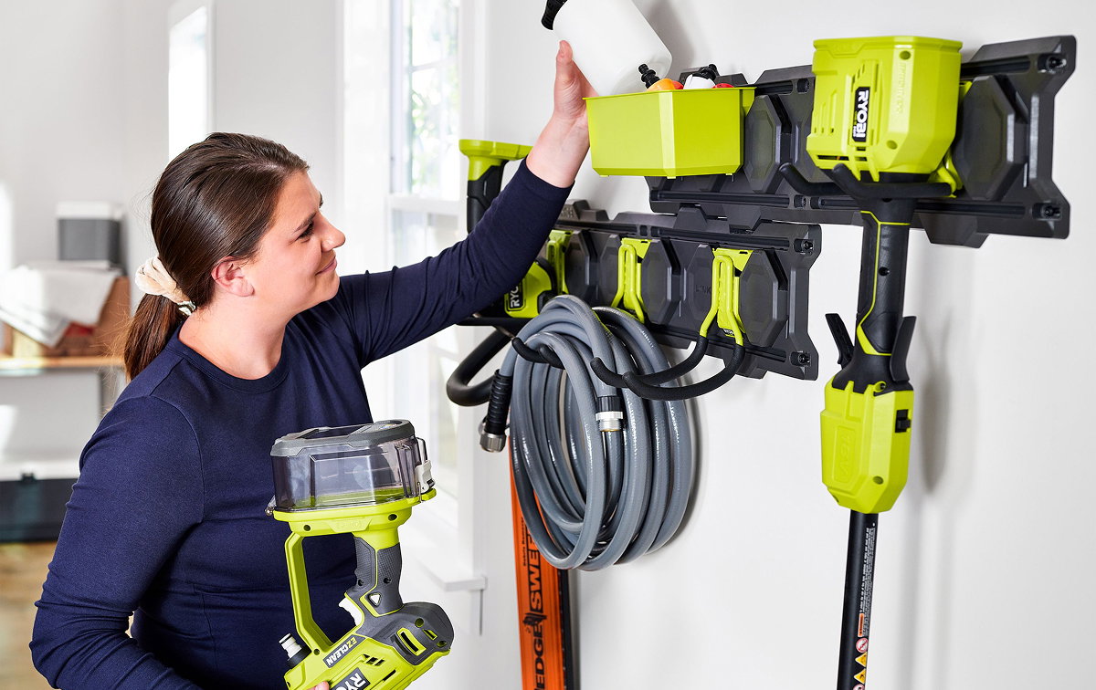 Woman in a navy shirt holding a RYOBI cordless vacuum while organizing tools and accessories on a RYOBI wall storage system in a garage