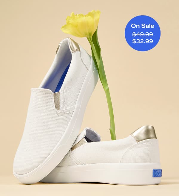 A pair of Pursuit Lurex Linen Slip-Ons in snow white/gold artfully positioned with a long-stemmed yellow flower.  A callout in the corner of the image notes that this sneaker was $49.99 and is now on sale for $32.99 for a limited time.