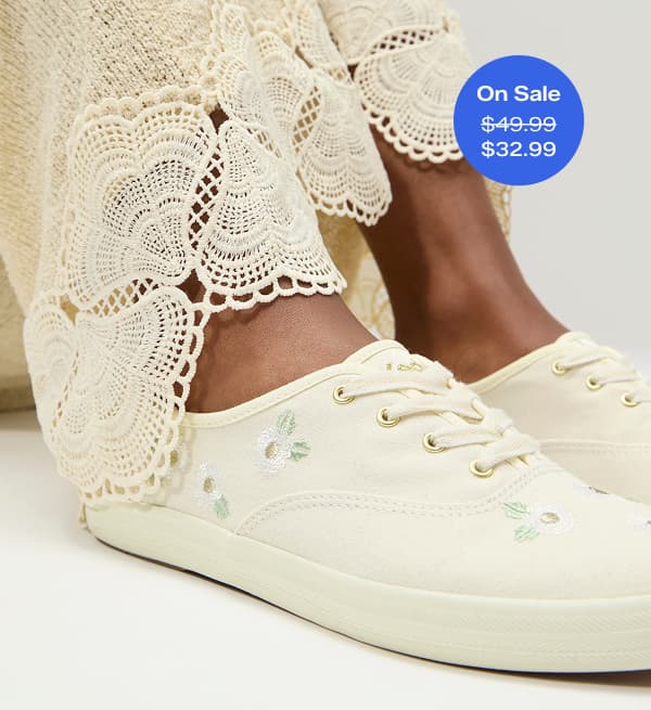 A closeup on a pair of Keds Champion Canvas Floral Embroidery Lace-Ups in egret shown on foot paired with cream-colored crochet pants. A callout in the corner of the image notes that this sneaker was $49.99 and is now on sale for $32.99 for a limited time.