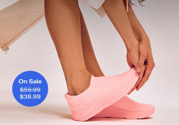 A woman wearing an ombre orange wind breaker jacket paired with the BlissMove Knit Ease Slip-Ons in neon coral. A callout in the corner of the image notes that this sneaker was $59.99 and is now on sale for $38.99 for a limited time. Text overlaid on the image says Spring Finale Style Edit.