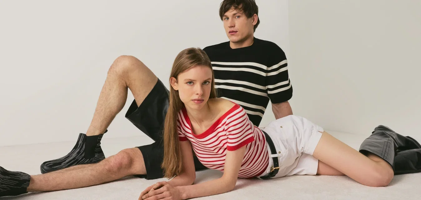 Jonge man en vrouw die poseren op de grond tegen een effen achtergrond, gekleed in gestreepte hemden, shorts en zwarte laarzen.
