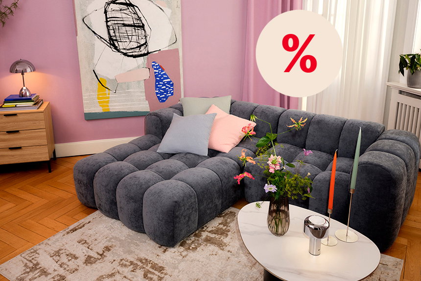 Wohnzimmer mit großem grauem Sofa, Blumen und Kerzen vor rosa Wand. Hinweis auf Rabatte.