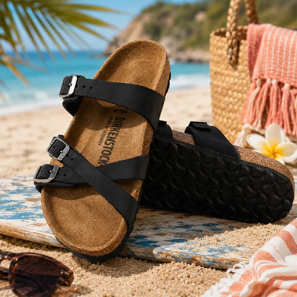 Women's Birkenstock sandals with black straps on sandy beach near ocean and palm trees