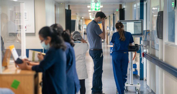 A view of a hospital corridor with various members of staff standing around - two look at a computer monitor.