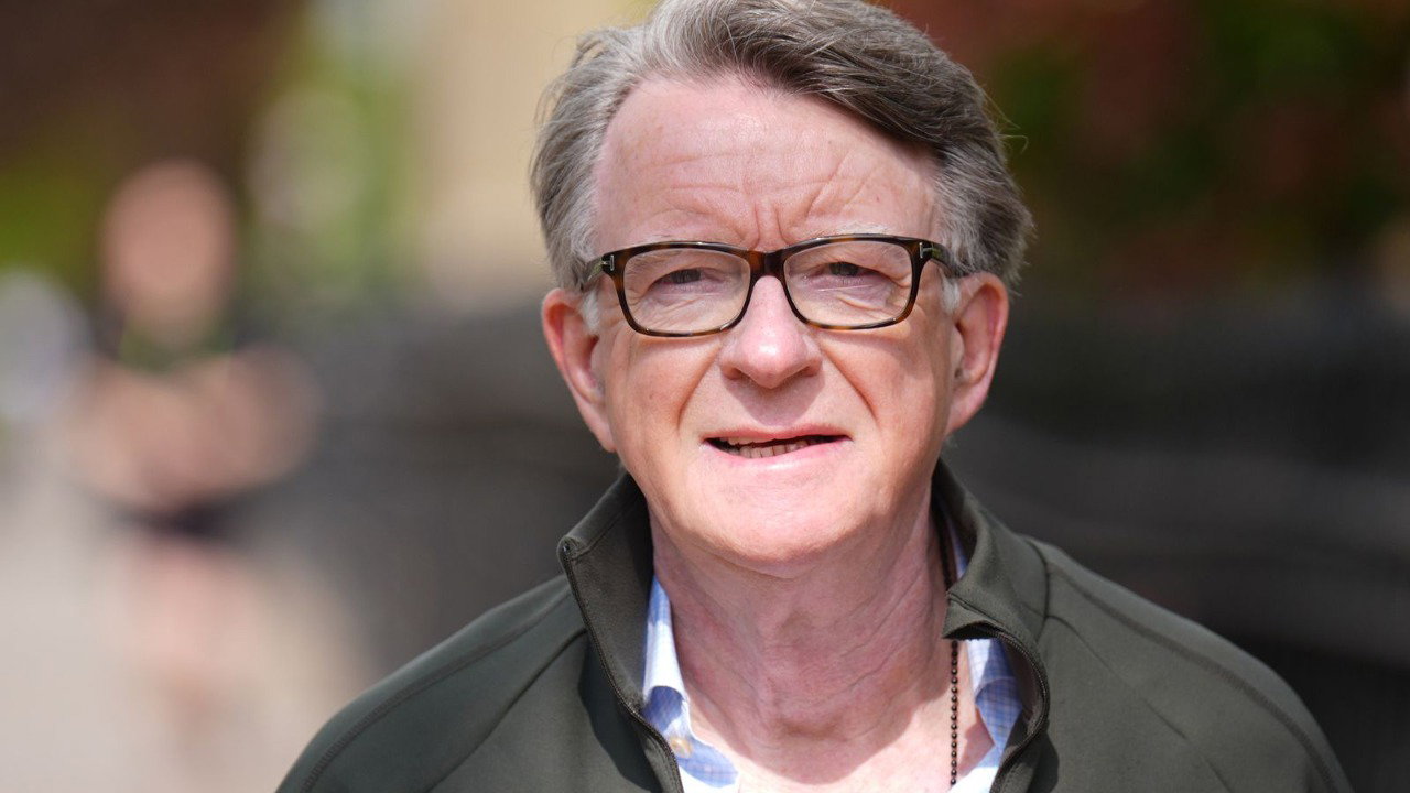 A headshot of Peter Mandelson, wearing  a green jacket and glasses. The outdoors background behind him is blurred.