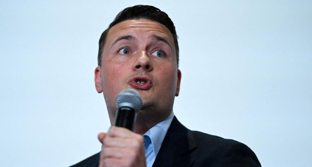A headshot of Wes Streeting from below as he speaks into a microphone. He is wearing a dark suit and is standing against a light blue background.
