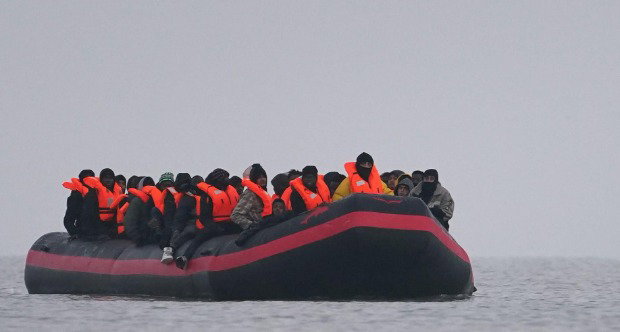 An inflatable boat out at sea, with dozens of people on board, wearing bright orange life-vests.