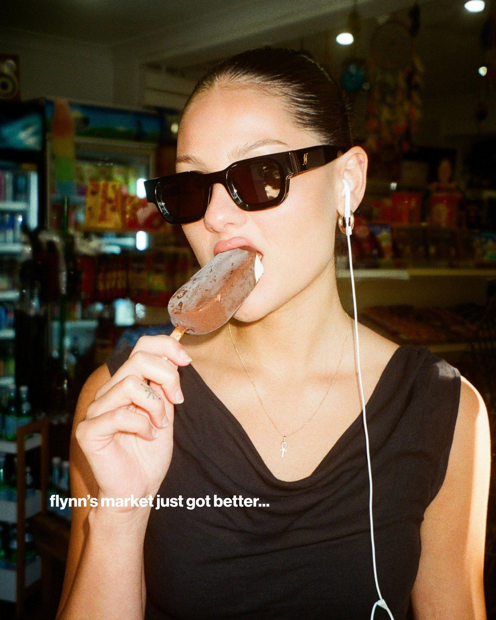 A medium shot captures a person wearing black sunglasses and a black top, holding a chocolate-covered popsicle to their mouth, with white earbuds in t...