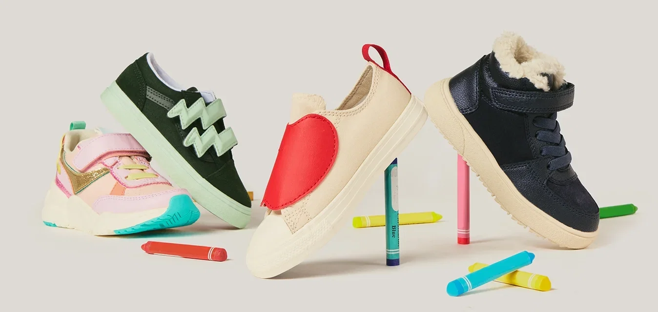 Quattro scarpe colorate per bambini in equilibrio su dei pastelli colorati, con altri pastelli sparsi su uno sfondo bianco.