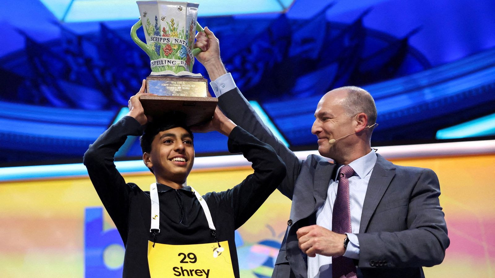 Shrey Parikh hold the spelling bee trophy above his head with both hands. He wears a black shirt and a yellow participant badge around his neck that reads ’29 Shrey’. A man wearing a grey suit assists Shrey in holding the trophy with one of his hands.
