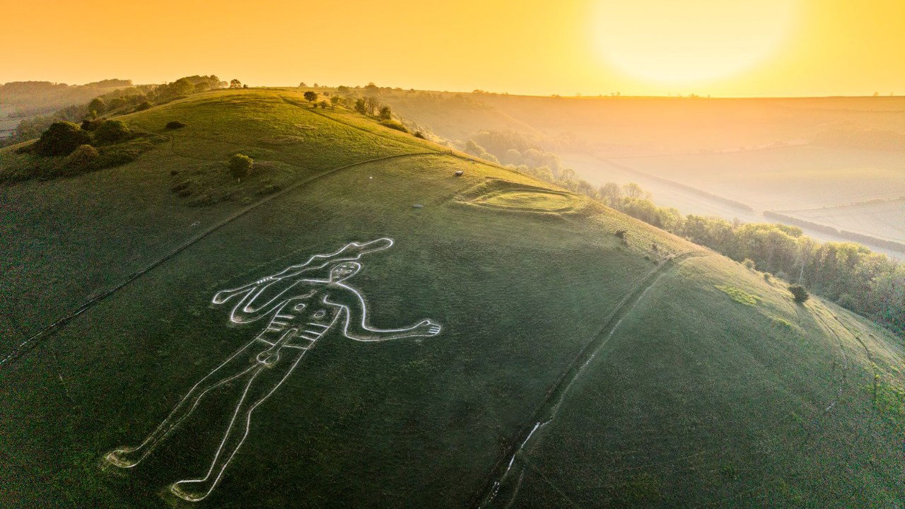 An aerial view of the Cerne Abbas Giant, which is etched onto a hillside in Cerne Abbas, Dorset. The large figure is naked and wields a club, detailed by its white chalk outline. The sun is setting on the horizon.