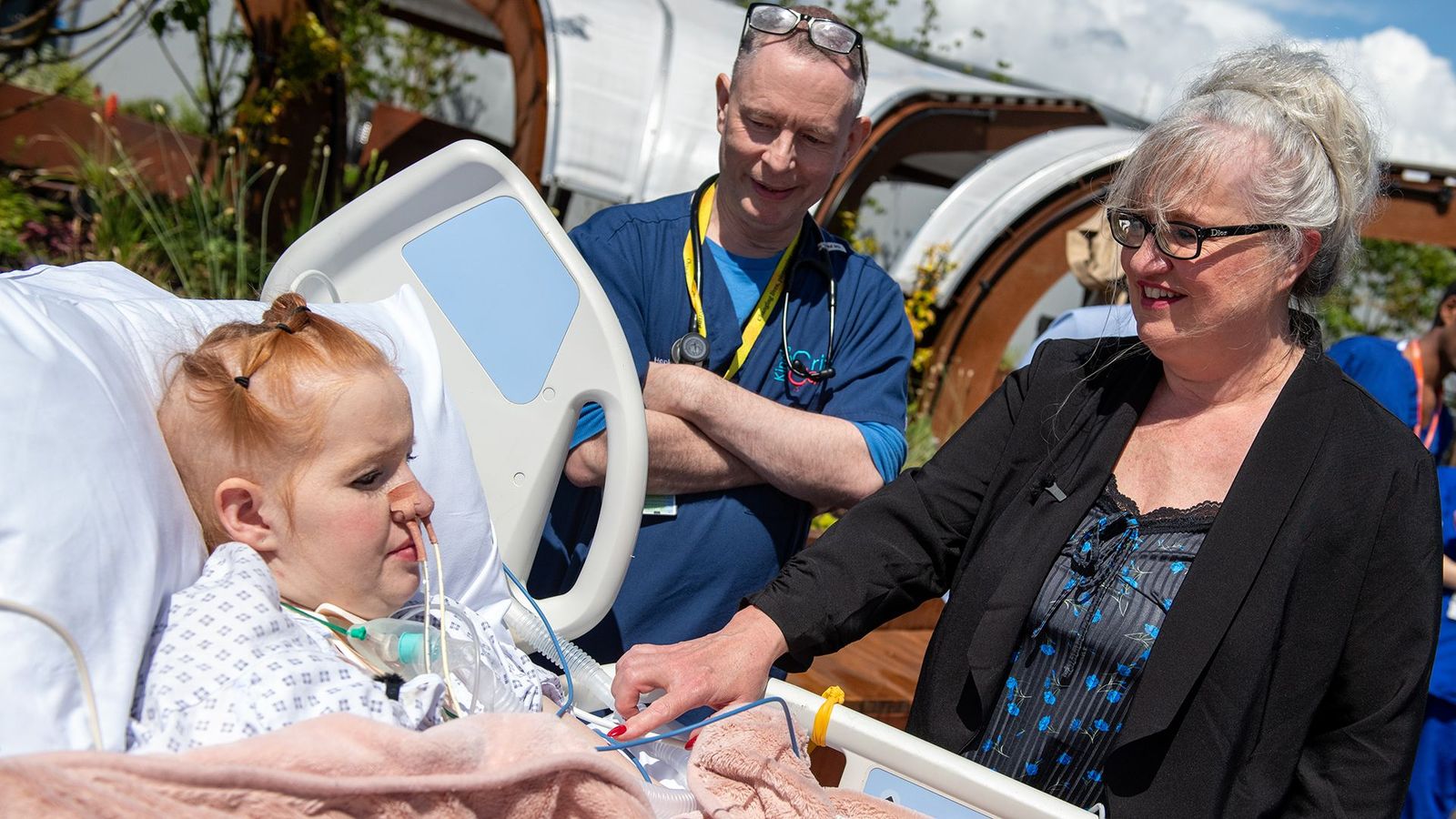 Hollie Allan, a patient at King’s College Hospital, sits in a hospital bed with multiple tubes attached to her. A male doctor with a stethoscope around his neck and an older woman with glasses stand next to her.