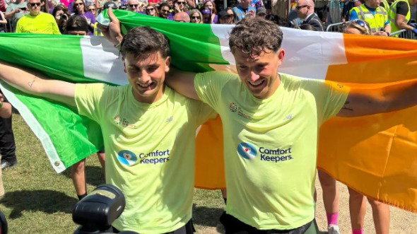 Brothers Jonathan and Cian Adams stand next to each other with their arms outstretched, both smiling and holding Ireland flags. They wear yellow t-shirts that read ‘Comfort Keepers’.