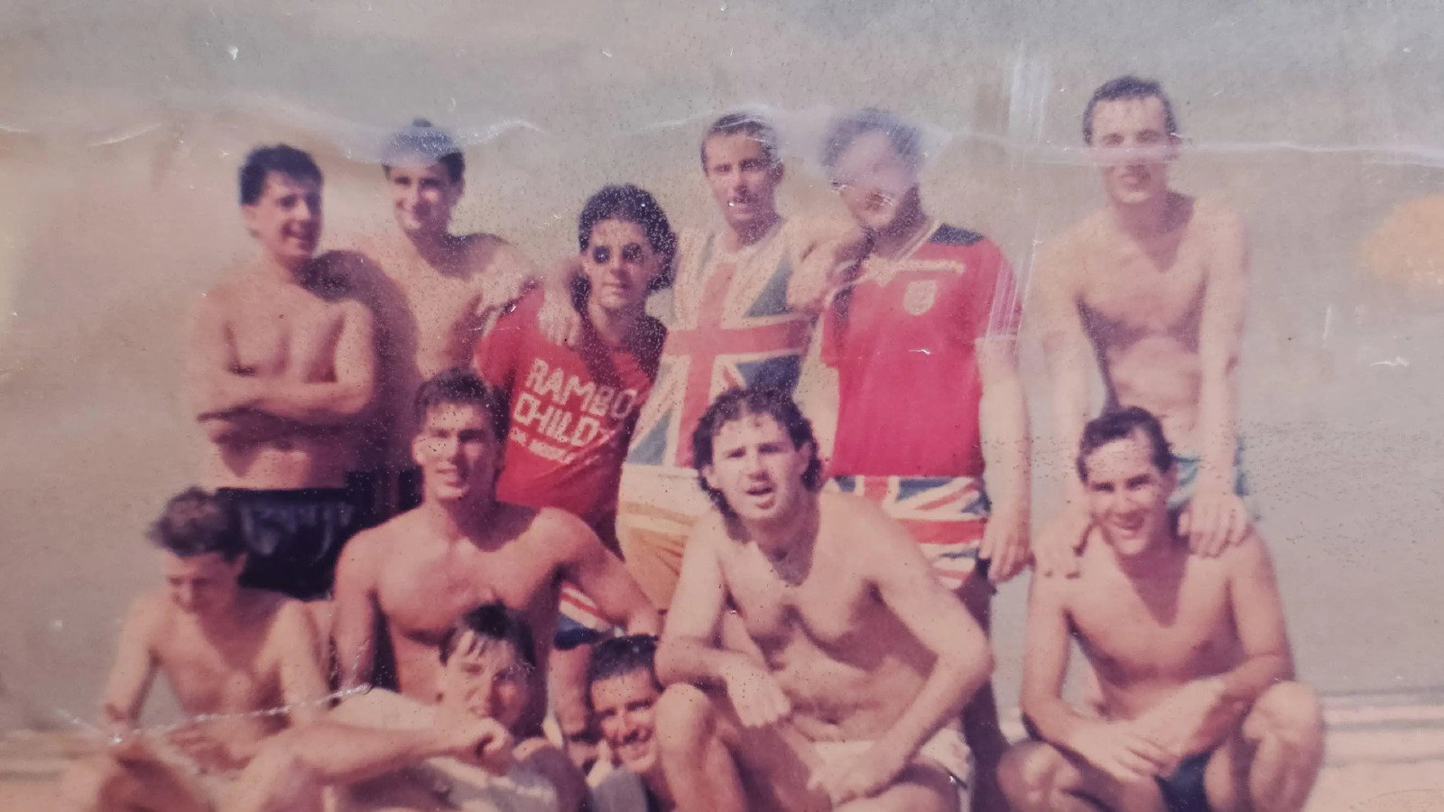 An old photograph of a group of friends with their arms around each other, some wearing shirts with the Union Jack flag on it.