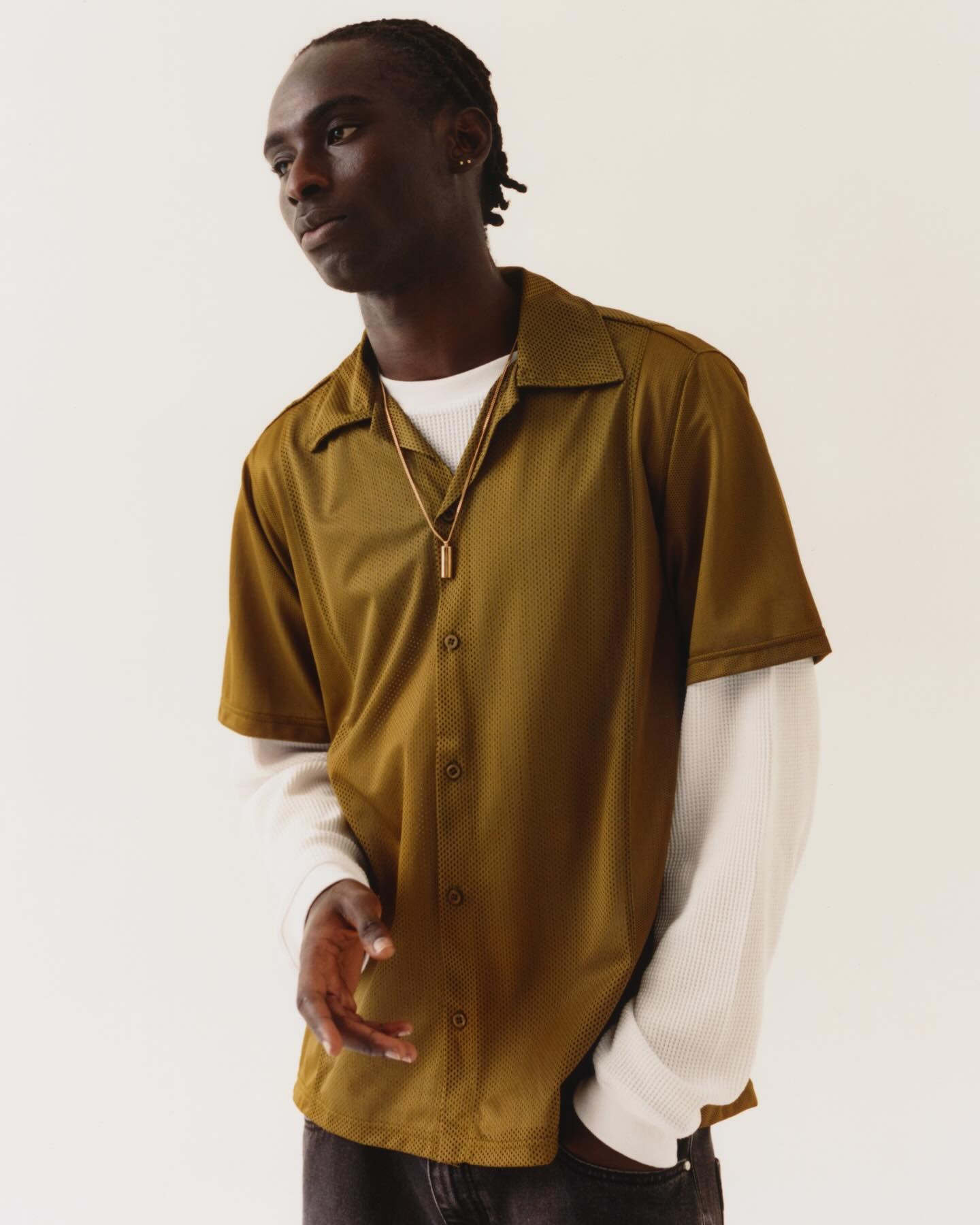Young man wearing olive-green short-sleeve button shirt over white long-sleeve top with gold pendant, standing against pale background