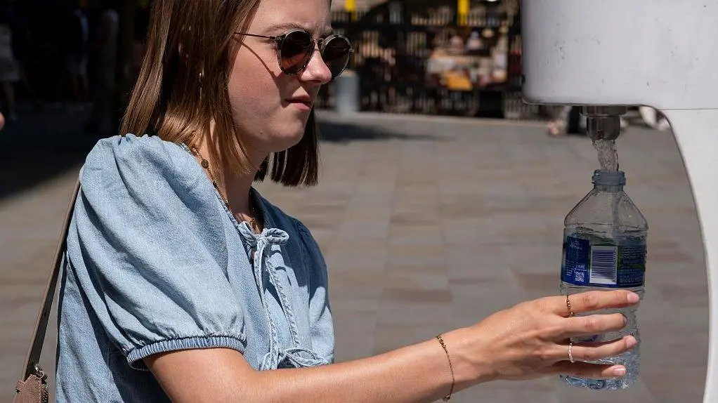 A woman wearing sunglasses stands outside on a sunny day. She is refilling her water bottle at an outdoor water dispenser unit.