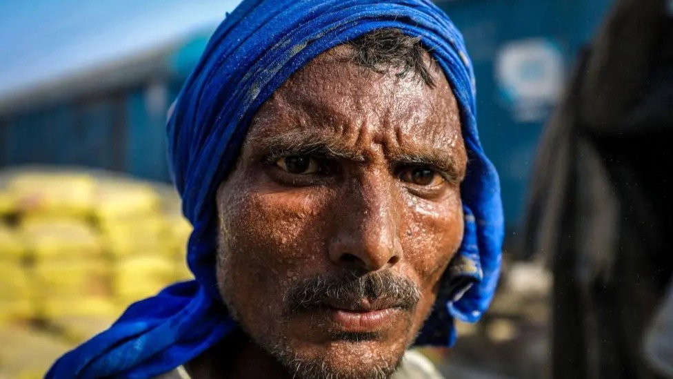 A close-up of a man standing outside in the sun with a blue scarf wrapped around his head.