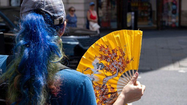 A woman with her blue hair in a ponytail and a cap is pictured from behind on a street, cooling herself with a yellow paper fan during hot weather.