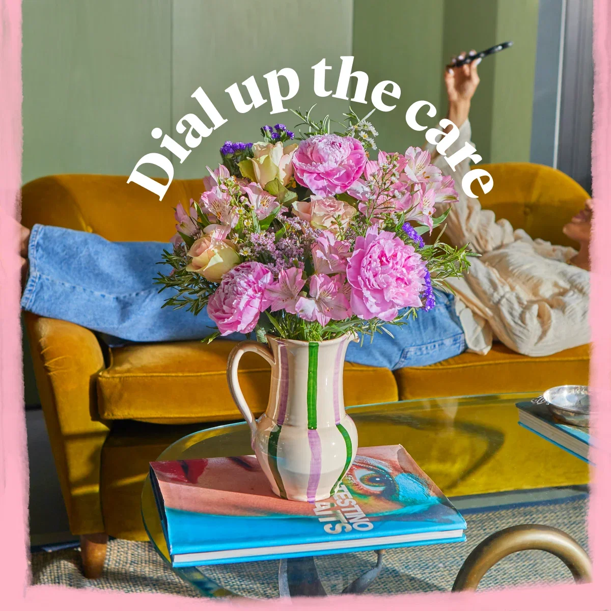 A vibrant pink and pastel bouquet arranged in a striped ceramic jug on a glass coffee table. The display features fluffy pink peonies, soft yellow roses, and delicate alstroemeria. In the background, a person relaxes on a mustard yellow velvet sofa while holding up a smartphone. Curved white text above the flowers reads ‘Dial up the care’.