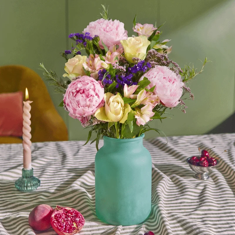 A vibrant summer bouquet featuring fluffy pink peonies, soft yellow roses, and deep purple statice beautifully arranged in a matte turquoise vase. The setting includes a striped green tablecloth, a lit pink twisted candle, and fresh sliced pomegranate. A soft pink oval sticker appears in the bottom right corner that reads ‘UK-grown peonies’.
