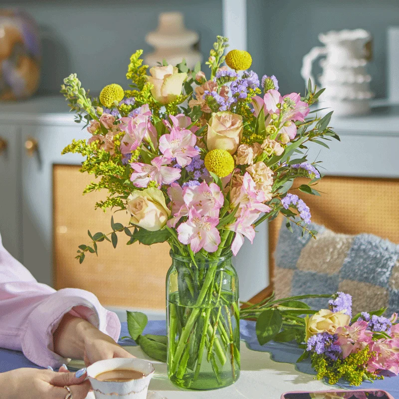 A bright bouquet arranged in a tinted green glass jar vase on a table. The display features spherical yellow craspedia, soft peach roses, pink alstroemeria, and light purple statice, with matching stems lying nearby ready to be added to the vase. A pink oval sticker appears in the bottom left corner that reads ‘Fabulous for ages’.