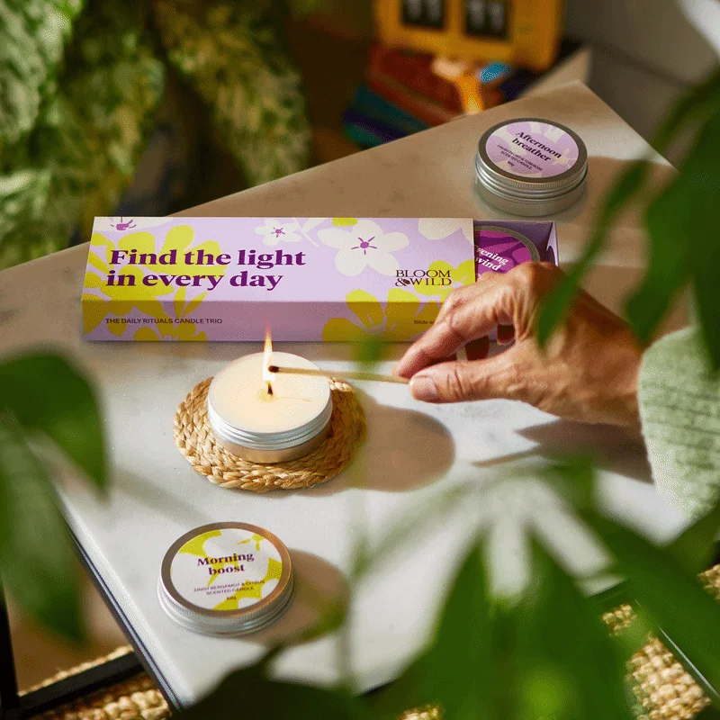 An overhead shot of a person lighting a small scented candle with a match on a marble tabletop. The candle sits alongside its lilac and yellow floral gift box, which reads ‘Find the light in every day: The Daily Rituals Candle Trio’. Individual candle tins are labelled ‘Morning boost’ and ‘Afternoon breather’. A pink oval sticker appears in the top left corner that reads ‘Exclusive to us’.