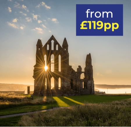 Ruins of an old abbey with sunlight shining through, set against a scenic landscape at sunset.