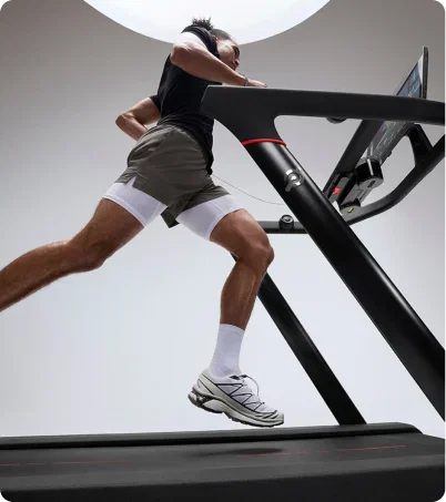 Man in black top and grey shorts running on Peloton Tread in bright studio