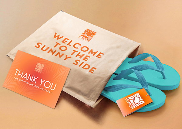 A brown mailing envelope that says “Welcome to the sunny side” is open on a table, with a pair of teal flip flops coming out of the top. An orange “Thank you” postcard sits against the mailer.