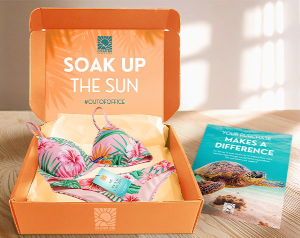 An open orange box printed with “Soak Up the Sun” on the inside top holds a colorful floral bikini, alongside a flyer featuring a sea turtle.