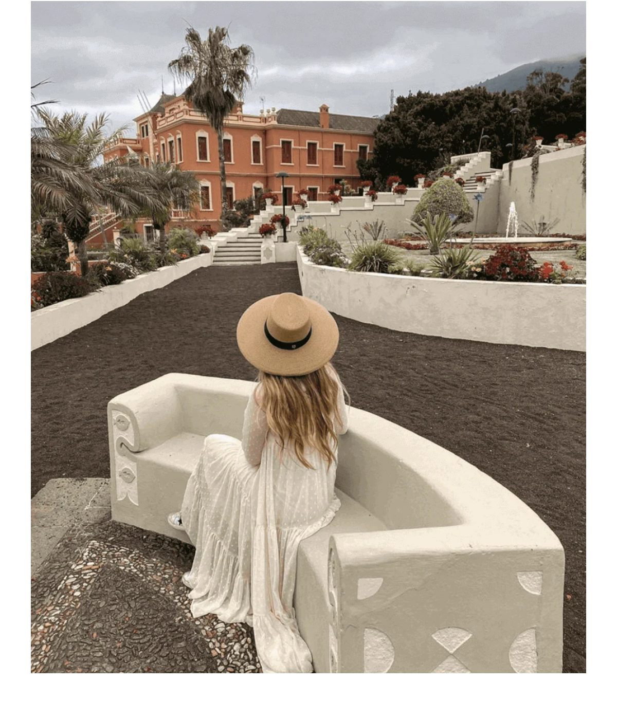 A woman in a white dress and hat sits on a curved stone bench in a garden setting.