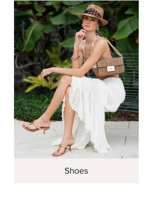 A woman in a white skirt and hat sits on a bench, holding a handbag, with greenery in the background.