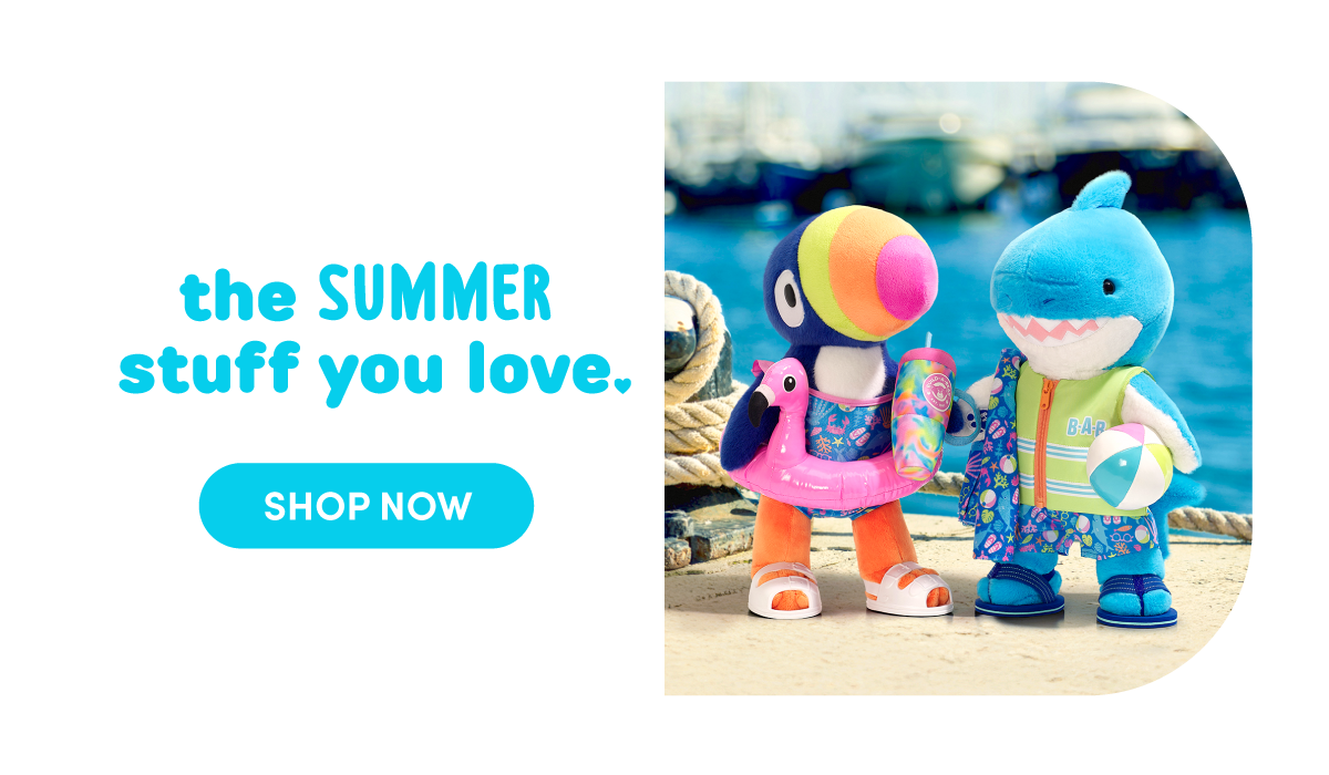 Marina setting with water, boats, and dock ropes on the right. A colorful toucan plush wearing a tropical swimsuit with a pink flamingo float stands beside a blue shark plush wearing a green vest and patterned swim trunks, holding a small beach ball. Text reads: the SUMMER stuff you love. SHOP NOW