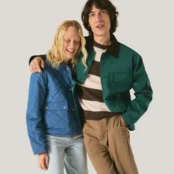 Dos jóvenes están juntos; uno lleva una chaqueta azul y jeans claros, el otro una chaqueta verde, camisa a rayas y pantalones marrones.